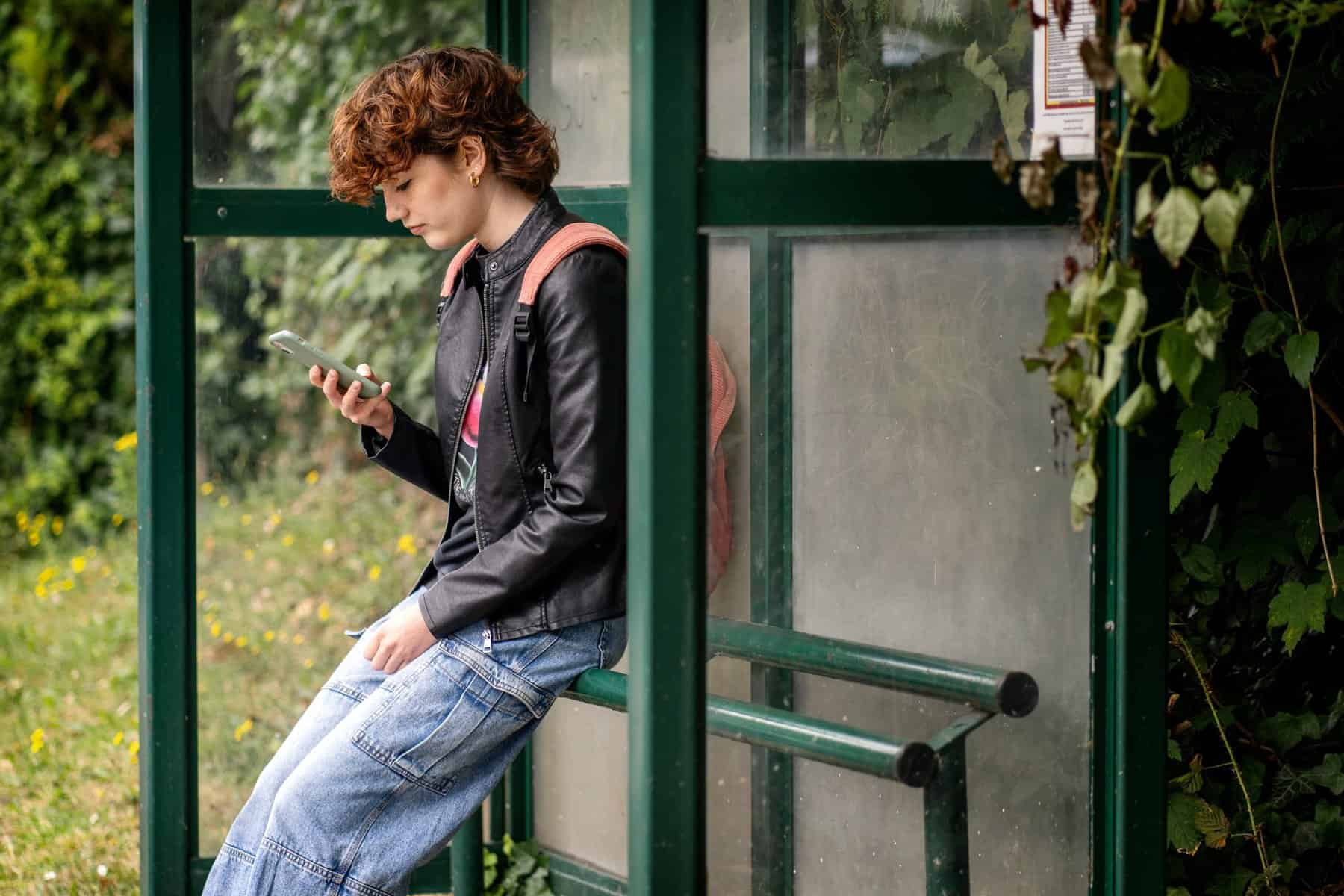 Teenage girl using smartphone at bus stop, promoting youth participation in digital access and safe technology.