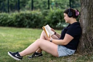 Young woman sitting against a tree, listening to music while reading a book outdoors.
