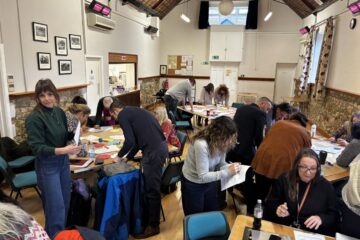 A room of people talking to each other and working at tables, learning about genuine safeguarding participation.
