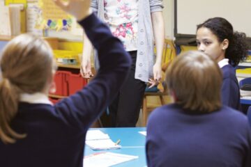 young pupils in a classroom holding up their hands, an example of pupil voice in schools